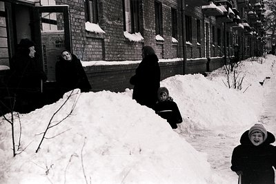 Three women and a child play in deep snow beside a Soviet-era brick apartment building, likely Eastern Europe, mid-20th centu...
