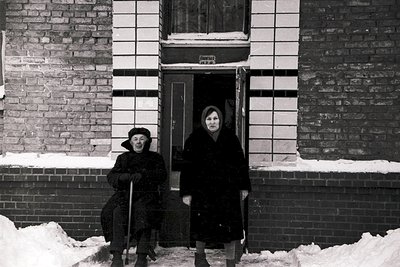 Black-and-white street scene featuring two elderly women in winter attire outside a brick apartment building. The seated woma...