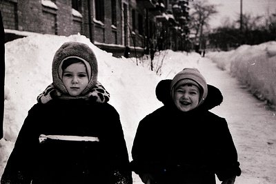 Two children in winter coats and knitted hats pose on a snow-covered path, mid-20th century. Snow blankets the ground and pil...