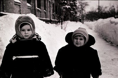 Two children in winter attire stand on a snow-covered path, bundled in heavy coats, knit hats, and scarves. The mid-20th cent...