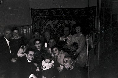 Vintage black-and-white group portrait in a dimly lit indoor setting, likely a home or communal space. Eight adults and two c...