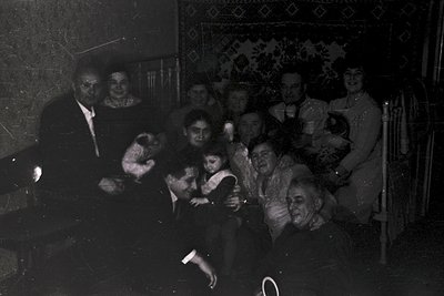 Vintage black-and-white family portrait in dim indoor lighting, likely 1940s–1950s. Group of 12–14 individuals seated on wood...