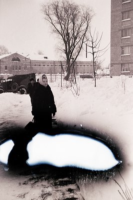 Winter street scene featuring a woman in a dark coat and headscarf standing beside a partially frozen puddle. Snow-covered ur...