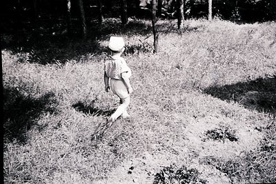 Child in mid-stride on overgrown grassy path, wearing shorts and a wide-brimmed hat, framed by dense forest foliage. Mid-20th...