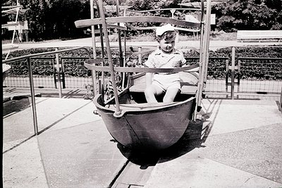 Mid-20th century child riding a vintage swing boat ride in a park. Black-and-white photo captures a child in a white cap and ...