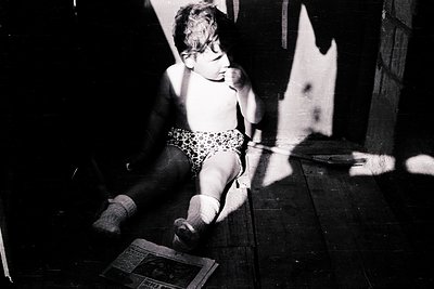 A young child sits cross-legged on a dark wooden floor, wearing patterned swim trunks and socks, with a book and shadow play ...