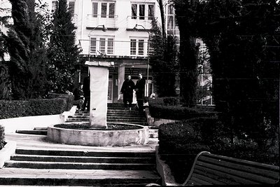 Mid-century courtyard with symmetrical stone steps leading to a grand entrance. Three men in suits converse near a central fo...