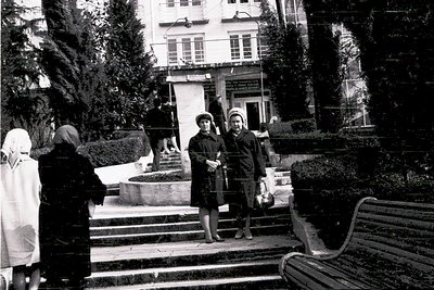 Mid-20th century urban courtyard scene featuring three women in winter attire—coats, hats, and scarves—posing near a fountain...