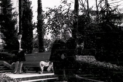 Black-and-white photo of a man in a forest clearing, seated on a wooden bench with a dog beside him. He wears a dark jacket a...