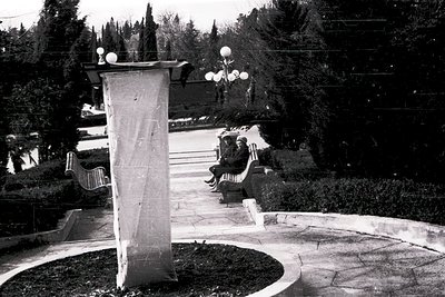 Two individuals sit on a stone bench in a landscaped park, framed by a tall, rectangular concrete monument with a speaker ato...