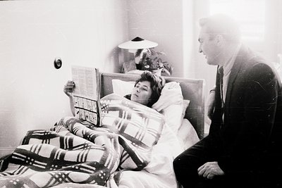 Mid-20th century hospital room scene: a man in suit reads a newspaper to a woman in bed, draped in plaid blanket. Wall-mounte...