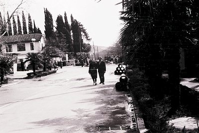 Black-and-white street scene featuring two uniformed individuals walking away from the camera, likely military or police. Res...