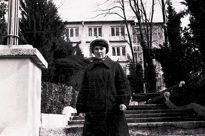 Black-and-white portrait of a woman in winter attire—coat, hat, and gloves—standing on a marble staircase beside a sculpted f...