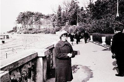 Mid-20th century seaside promenade with concrete steps and stone barriers. Woman in 1960s-style coat and hat poses near water...