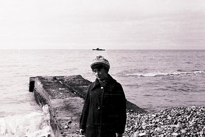 Mid-20th century seaside portrait: individual in winter attire (beanie, coat) stands on rocky pier by choppy waters. Distant ...