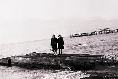 Two women in early 20th-century attire stand on a concrete pier by the sea, one holding a bag. Industrial fishing nets and wo...