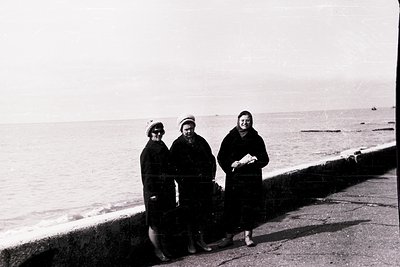 Three women in early-to-mid 20th-century attire pose by a seaside promenade, likely Eastern European. Black-and-white photo c...