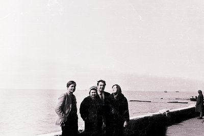 Black-and-white seaside group portrait on a stone pier, likely 1960s–1970s. Four individuals in winter coats pose near calm w...