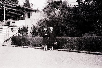 Two young girls in early 20th-century attire pose on a paved roadside, one holding a small handbag. The girl on the left wear...