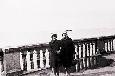 Two individuals in mid-20th-century attire pose on a seaside promenade with ornate balustrade, likely . Woman wears a fitted ...