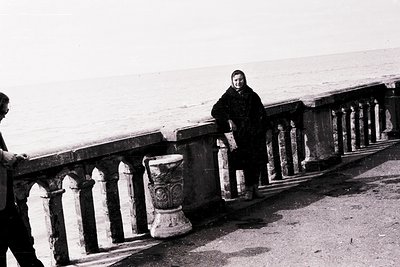 Black-and-white seaside scene featuring a woman in a heavy coat leaning on ornate balustrade, likely –. Wavy, stormy sea in b...