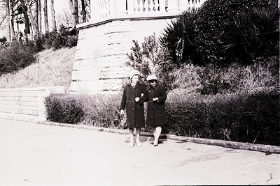 Two women in 1950s-style coats and hats pose on a sunlit seaside promenade, flanked by stone steps and lush greenery. Classic...