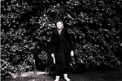 Black-and-white portrait of a child in winter attire—hooded coat, scarf, and boots—standing among dense evergreen foliage. Th...