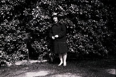 Black-and-white portrait of a woman in a structured coat and hat, standing outdoors among dense foliage. Her pose suggests mi...