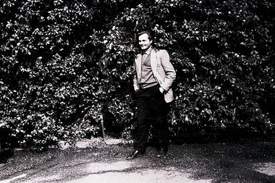Portrait of a man in a light-colored suit and tie, standing confidently in a wooded area with dense foliage. The monochrome s...