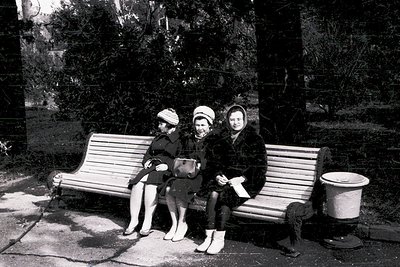 Three women in 1960s-era winter attire sit on a wooden bench in an urban park. Dark coats, hats, and stockings suggest cold w...