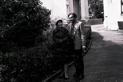 Black-and-white portrait of a man and woman in mid-20th-century attire, posing outdoors near a building with arched doorway. ...