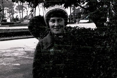 Vintage black-and-white portrait of a woman in winter attire—fur-trimmed coat, hat with a feather, and gloves—posing outdoors...