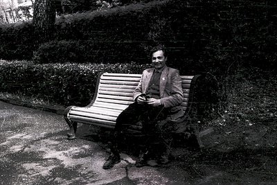 Mid-century man in tailored suit and bowtie sits on a wooden bench in a park, holding a small device (likely a walkie-talkie ...