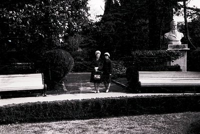 Two women in 1960s-era attire walk through a landscaped park, one carrying a bag. The woman on the left wears a light jacket ...