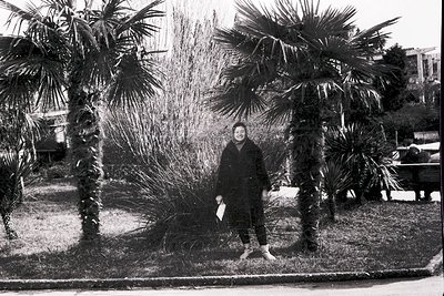 Black-and-white street portrait of a person standing beside tall palm trees in an urban park, holding a folded paper. Mid-cen...