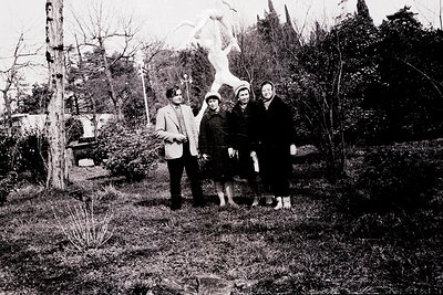 Four individuals pose outdoors near a stone cross in a rural setting, mid-20th century. Formal attire suggests a special occa...