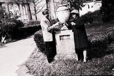 Two individuals pose outdoors in a formal setting, likely mid-20th century. The man, in a light jacket and dark trousers, han...