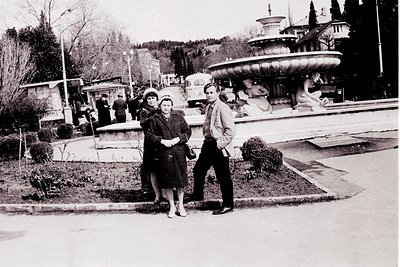 Black-and-white photo of three centrally posed individuals in a public square with a prominent Art Nouveau-style fountain. Me...