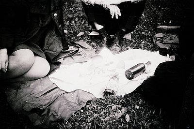 Black-and-white photo of three individuals seated on grass, examining a rolled map or document spread on a cloth. A dark bott...