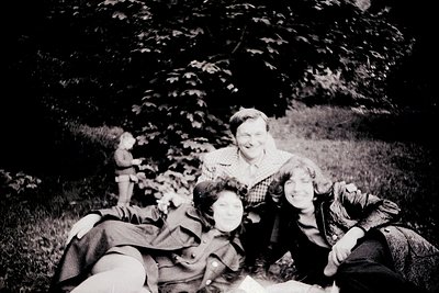 Vintage black-and-white photo of three individuals lounging on grass under leafy trees, mid-20th century. Central figure wear...