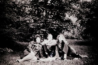 Vintage black-and-white portrait of three individuals seated closely in a wooded area, likely mid-20th century. Natural light...
