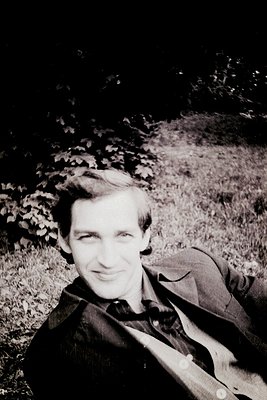 Black-and-white portrait of a man in a relaxed pose on grass, leaning against a tree trunk. His short hair and dark suit sugg...