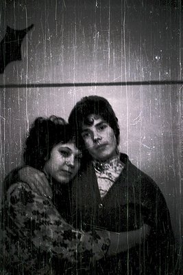 Two individuals stand under a shower curtain in a vintage-style bathroom, captured in black-and-white. The woman wears a flor...