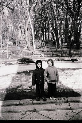 Two children pose on a cracked concrete path in a winter forest, bundled in 1950s-style winter coats and scarves. The girl we...
