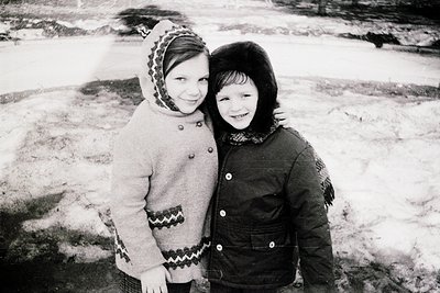 Two children pose closely in winter attire, likely mid-20th century. Girl wears a knitted coat with geometric pattern and hea...