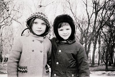 Two children pose outdoors in winter attire, likely mid-20th century (1950s–1970s). Girl wears a knitted hat with floral trim...