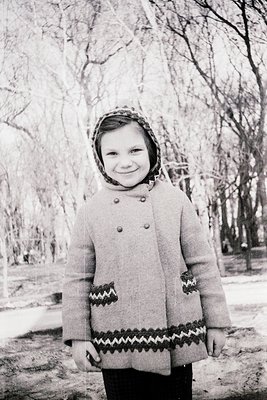 Black-and-white portrait of a young girl in winter attire: knitted coat with zigzag trim, buttoned cuffs, and a headscarf. Le...