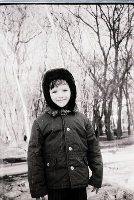 Mid-century winter portrait of a child in a buttoned-up quilted jacket and fur-lined hood, standing on a snow-dusted path. Ba...