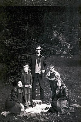 Family portrait in a forested setting, mid-20th century. Five individuals pose outdoors on a blanket, surrounded by dense fol...