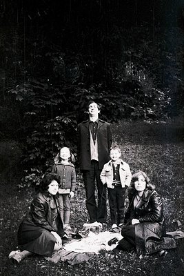 Family portrait in a wooded forest, mid-20th century. Five individuals pose on a blanket: two adults seated, three children s...